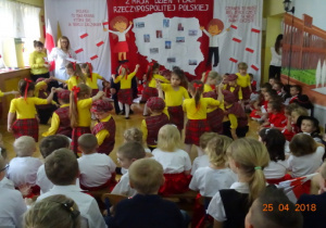 Na tle dekoracji z mapą Polski i dziećmi z chorągiewkami przedszkolaki tańczą w parach. Na krzesełkach siedzą dzieci, które oglądają taniec.